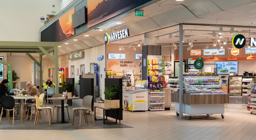 Narvesen kiosk på flyplass med mat, drikke og seter til kundar. Her ser vi fleire personar som sit og ventar ved bord, omgitt av grønne planter. Skilt for "Arrivals" heng i taket
