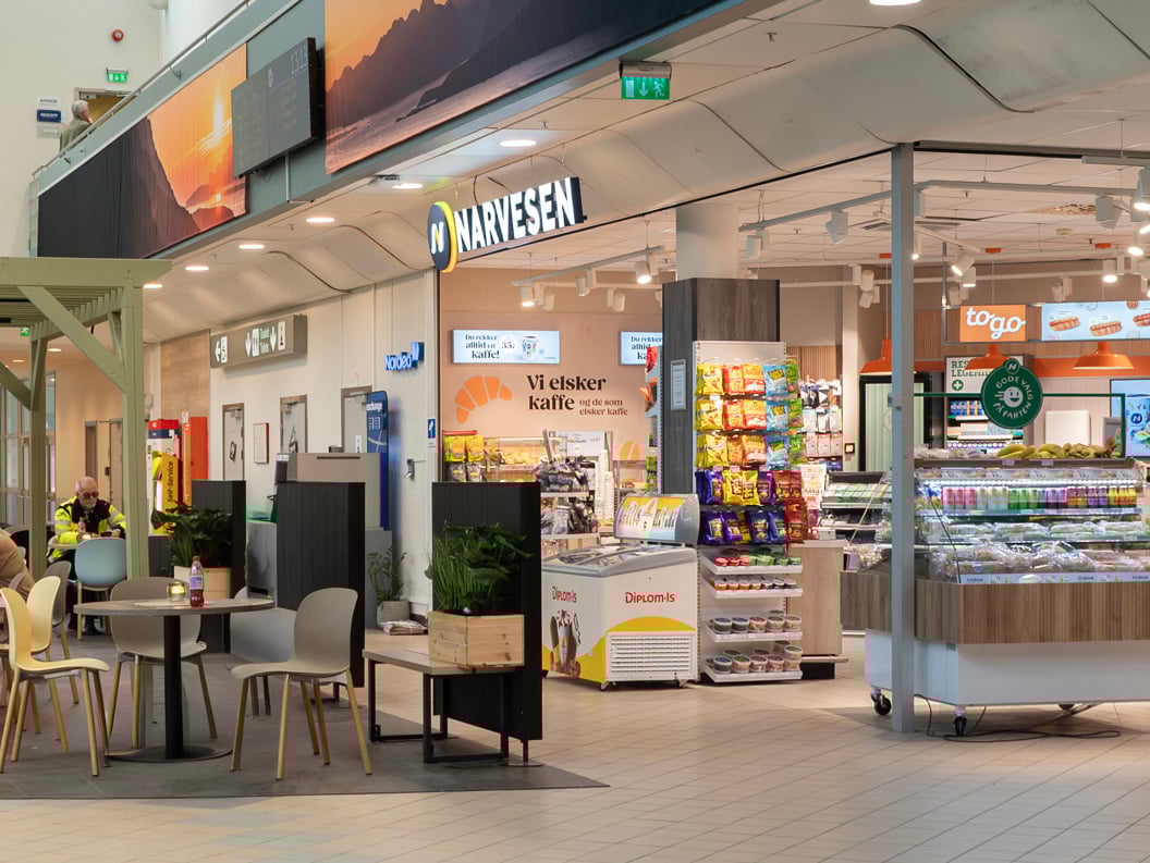 Moderne flyplasskiosk og kafé med sittende kunder, med Narvesen-branding, produktdisplayer og ankomstskilt