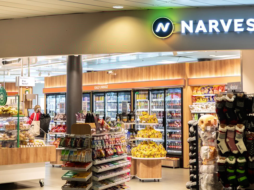 Narvesen store interior at an airport, featuring a variety of snacks, beverages, magazines, and travel essentials, with people browsing near seating areas