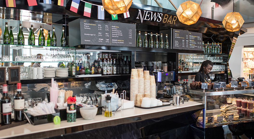 Modern café bar interior with drink selections, sandwich and pastry display, and a person serving behind the counter at News Bar, decorated with international flags and warm lighting