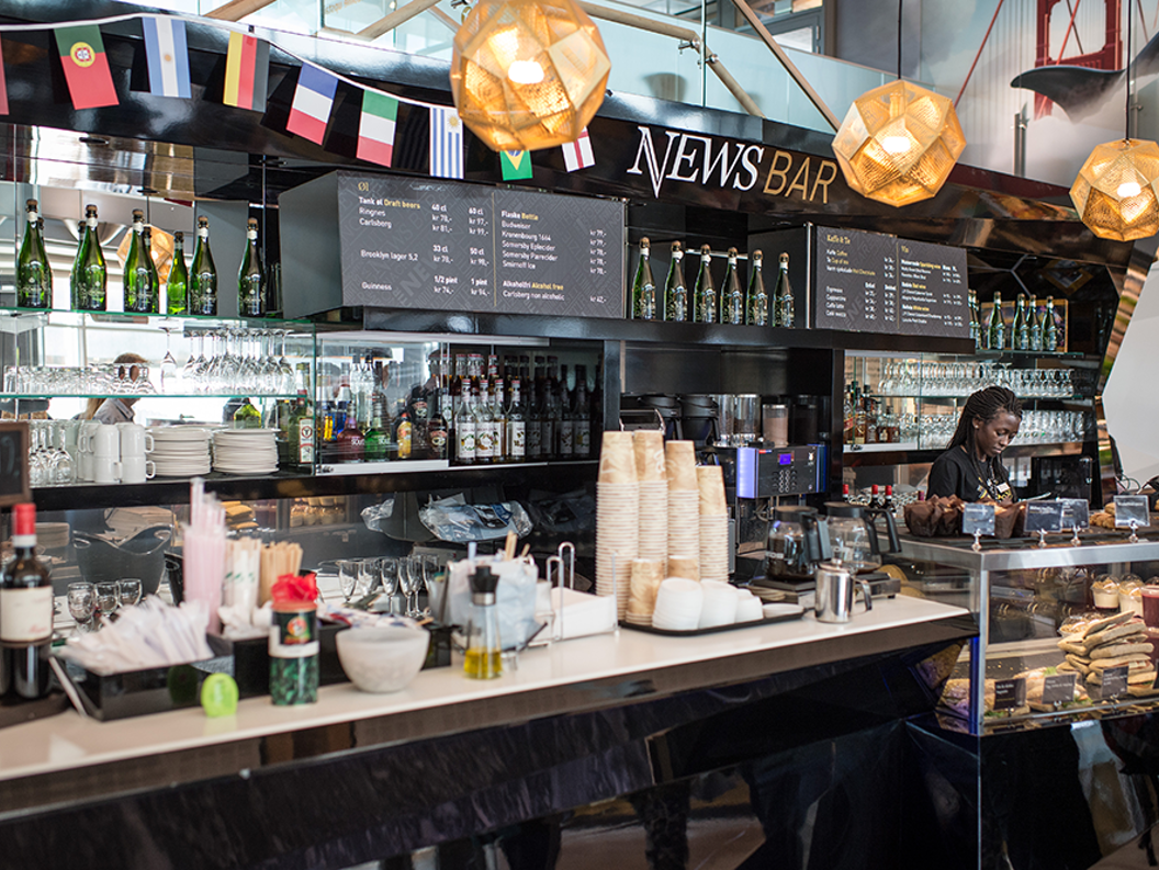 Moderne kafébarinteriør med utvalg av drikkevarer, smørbrød- og bakevaredisplay og en person som serverer bak disken på News Bar, dekorert med internasjonale flagg og varm belysning