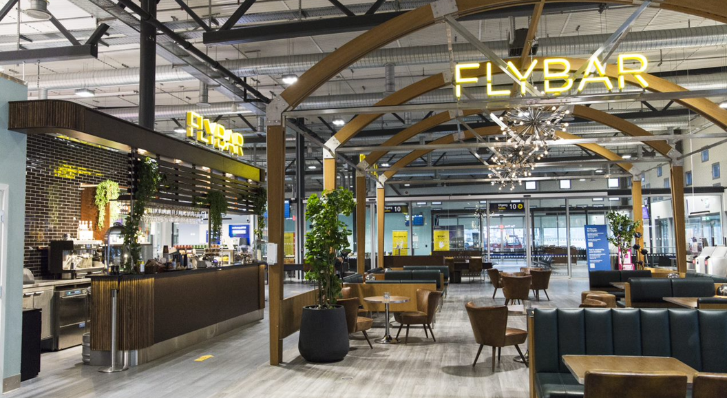 Modern airport lounge with Flybar signage, featuring stylish seating, a sleek bar area, decorative lighting, and plants in a spacious, industrial-themed setting