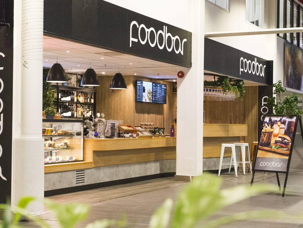 Modern food bar interior with wooden counters, black pendant lighting, and a digital menu display. A-frame sign showcasing a burger meal in the foreground
