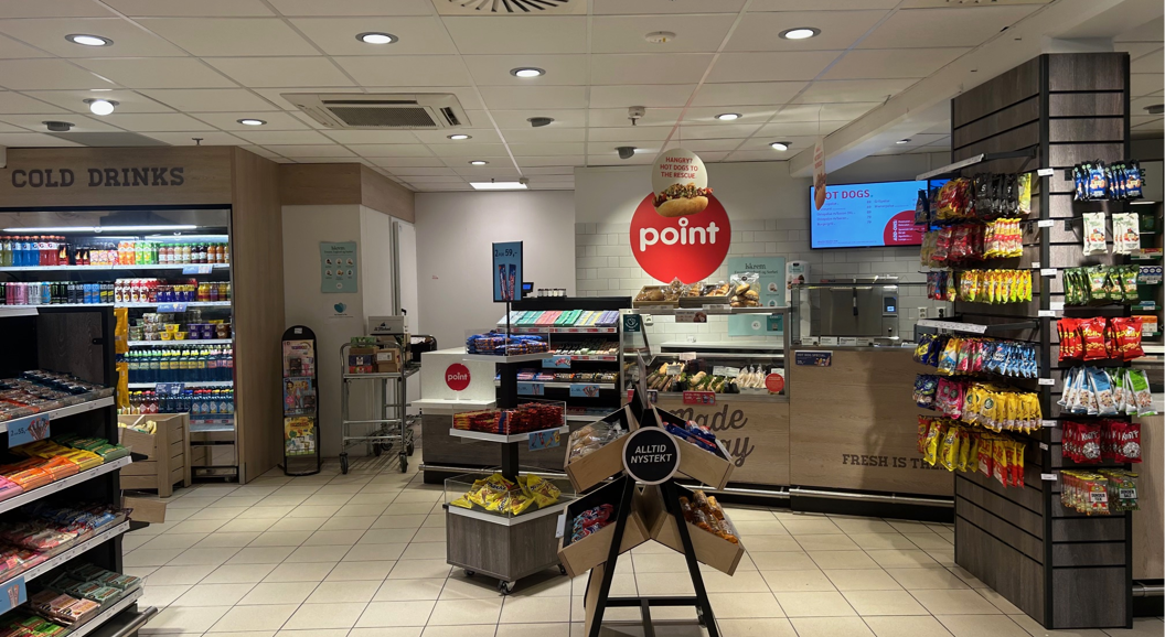 Inngang til en dagligvarebutikk med fruktutstilling, magasiner, kalde drikker og en delikatesseavdeling med skiltet "Laget i dag". Ulike produkter er tilgjengelige for kundene