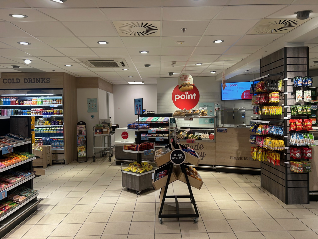 Inngang til en dagligvarebutikk med fruktutstilling, magasiner, kalde drikker og en delikatesseavdeling med skiltet "Laget i dag". Ulike produkter er tilgjengelige for kundene