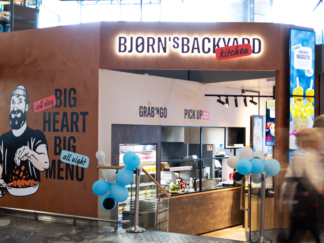 Bjorn's Backyard Kitchen restaurant entrance in a shopping mall, featuring a mural of a bearded man holding food, with slogans "Big Heart Big Menu" and "Grab 'N Go, Pick Up