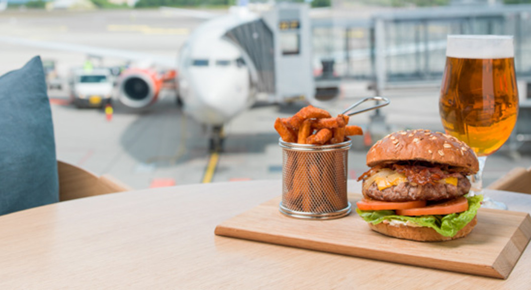 Hamburgar med salat og tomat, søtpotetfries og eit glas øl på eit bord på ein flyplass, med eit fly i bakgrunnen