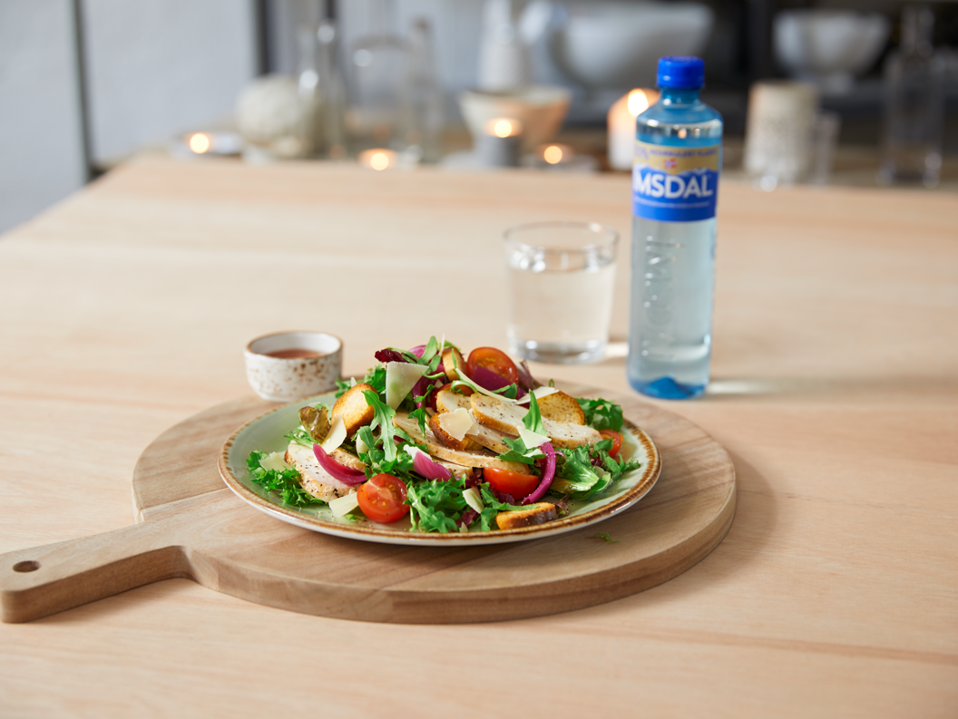 Fresh mixed salad with cherry tomatoes, croutons, and greens on a wooden board, accompanied by a small dressing cup, glass of water, and bottled water in a well-lit dining setting
