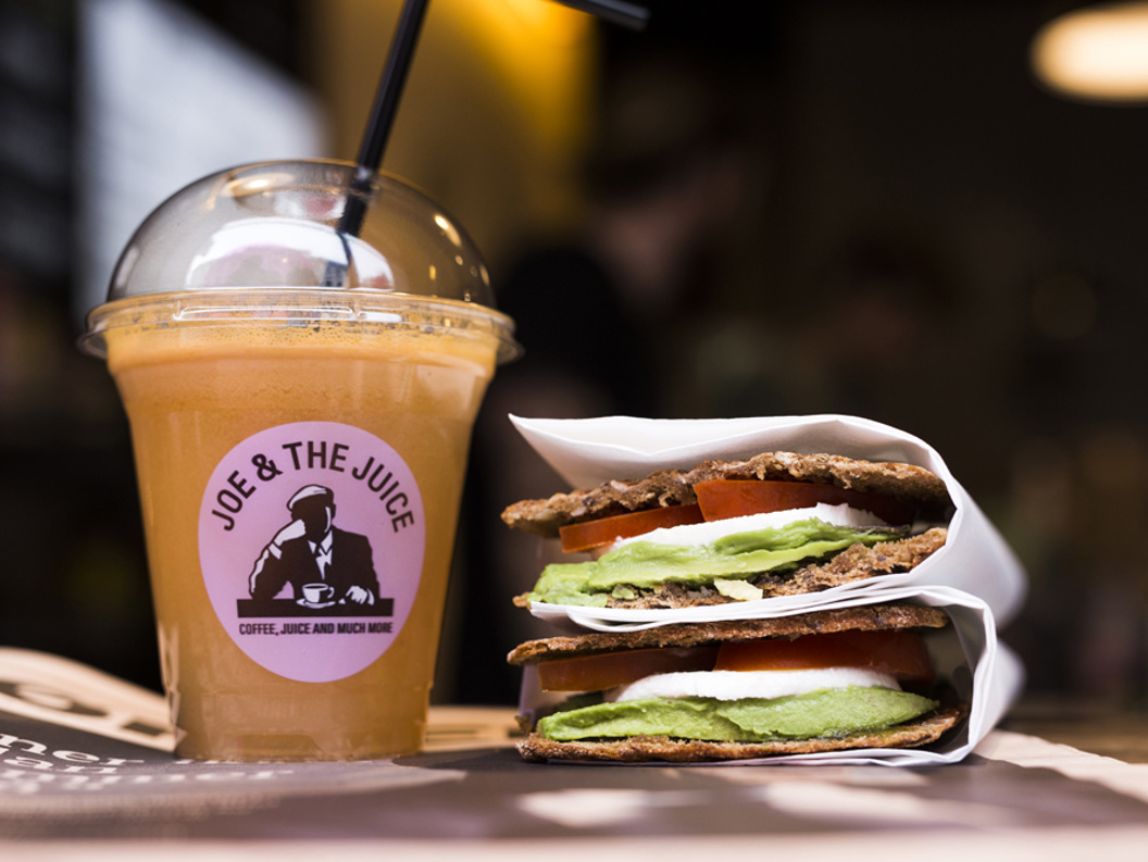 Freshly made sandwich with avocado and tomato paired with a frothy orange juice in a Joe & The Juice branded cup