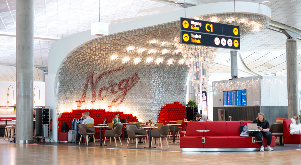 Interiør ved norsk flyplasslounge med ordet "Norge" i bakgrunnen, raud sofa, reisande som sit og kviler, og skilt som viser veg til utgang C1 og toalett