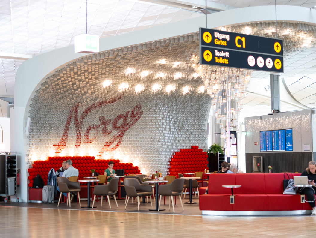 Norsk flyplasslounge med sittegruppe, dekorativ veggdesign med 'Norge', retningsskilt for porter og toaletter, og passasjerer som jobber på bærbare datamaskiner