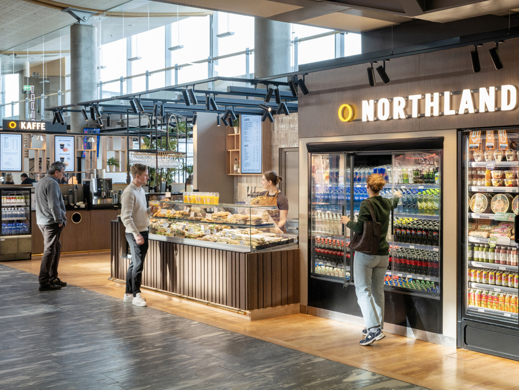 Kiosk i flyplass med folk som kjøper mat og drikke frå diskar og kjøleskap hjå Northland, moderne interiør i lyse omgjevnader