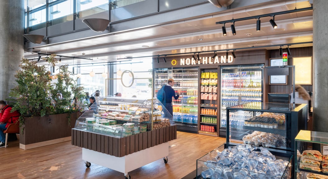 Modern airport convenience store interior with refrigerated drinks, snacks, and fresh food display at Northland shop. Shoppers browse aisles in a well-lit, open space with wooden flooring and contemporary design elements