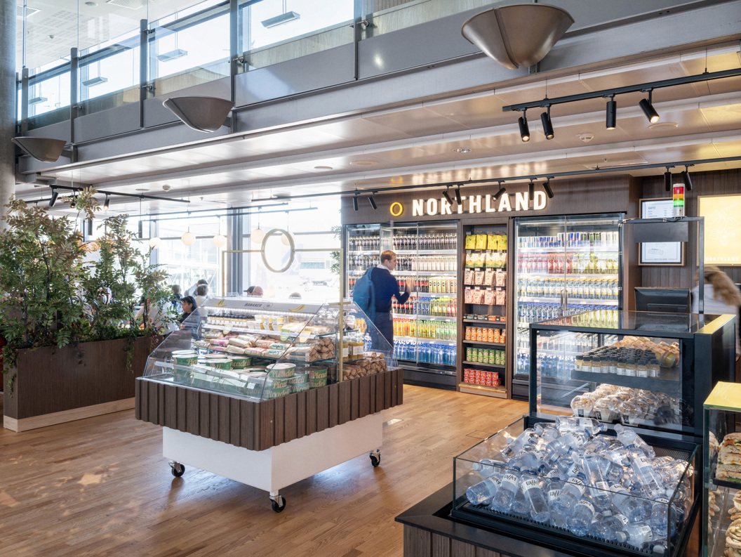 Modern airport convenience store interior with refrigerated drinks, snacks, and fresh food display at Northland shop. Shoppers browse aisles in a well-lit, open space with wooden flooring and contemporary design elements
