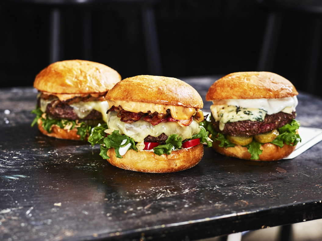 Tre gourmetburgere med ulike toppinger, servert på et rustikt svart bord, som fremhever saftige biffburgere, frisk salat og smeltet ost