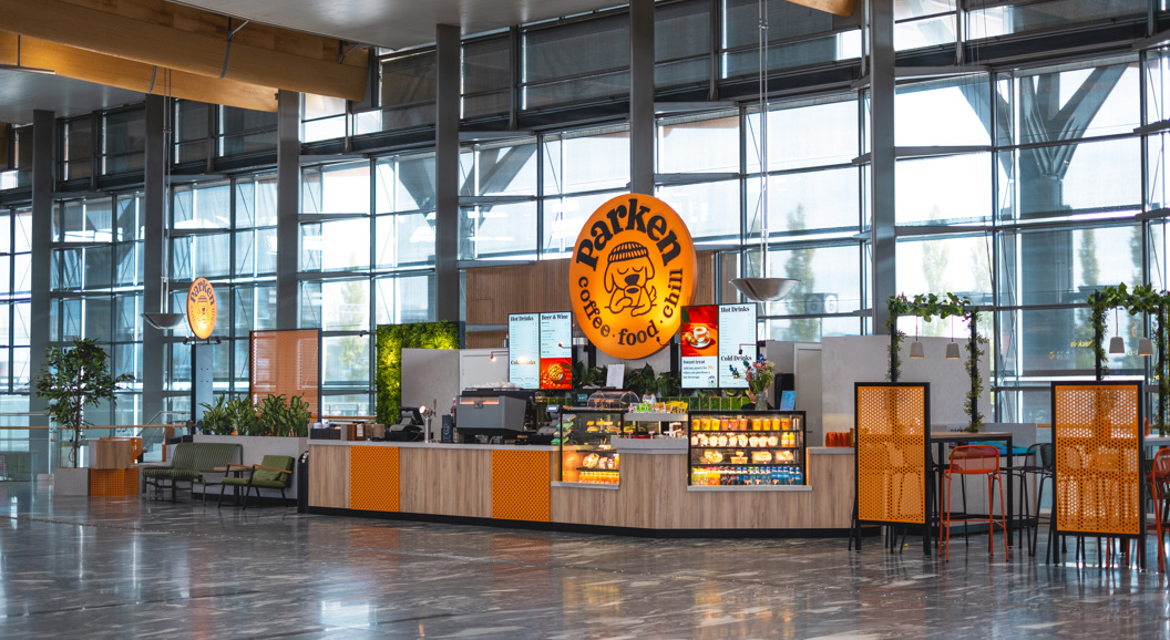 Kafé i moderne terminalbygg med store glasvegger, skiltet "Barken" synleg, og fargerikt interiør