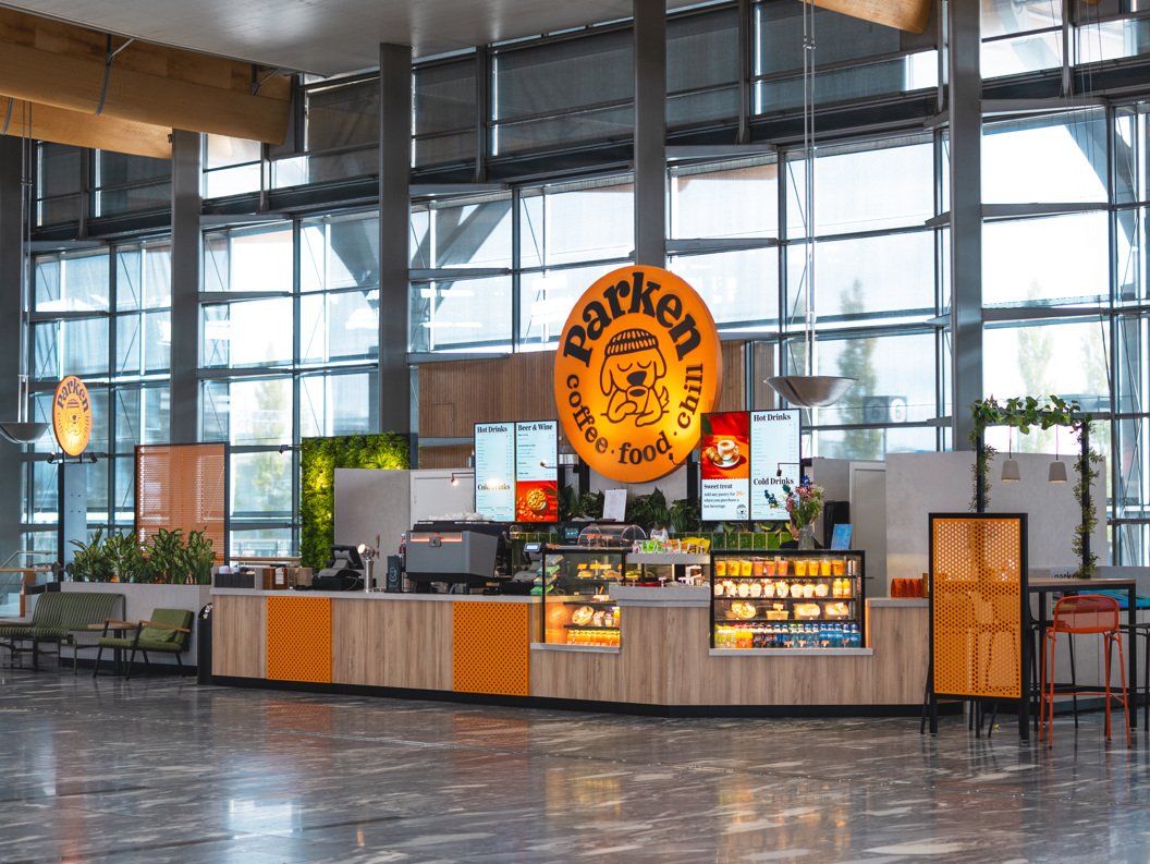Kafé i moderne terminalbygg med store glasvegger, skiltet "Barken" synleg, og fargerikt interiør
