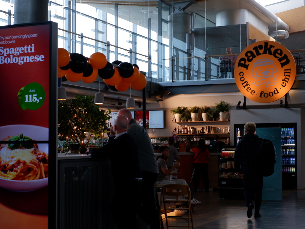 Interiør i en flyplasskafé med et skilt som reklamerer for spaghetti bolognese for 115 valutaenheter; kaféen heter Parken og har en avslappet atmosfære med sitteplasser og planter