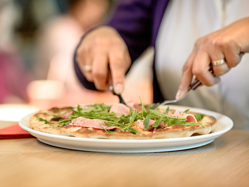 Person som skjærer en pizza toppet med ruccola og prosciutto på en hvit tallerken.