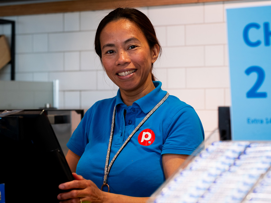 Smiling cashier in a blue uniform at a convenience store counter, with a promotional sign in the background