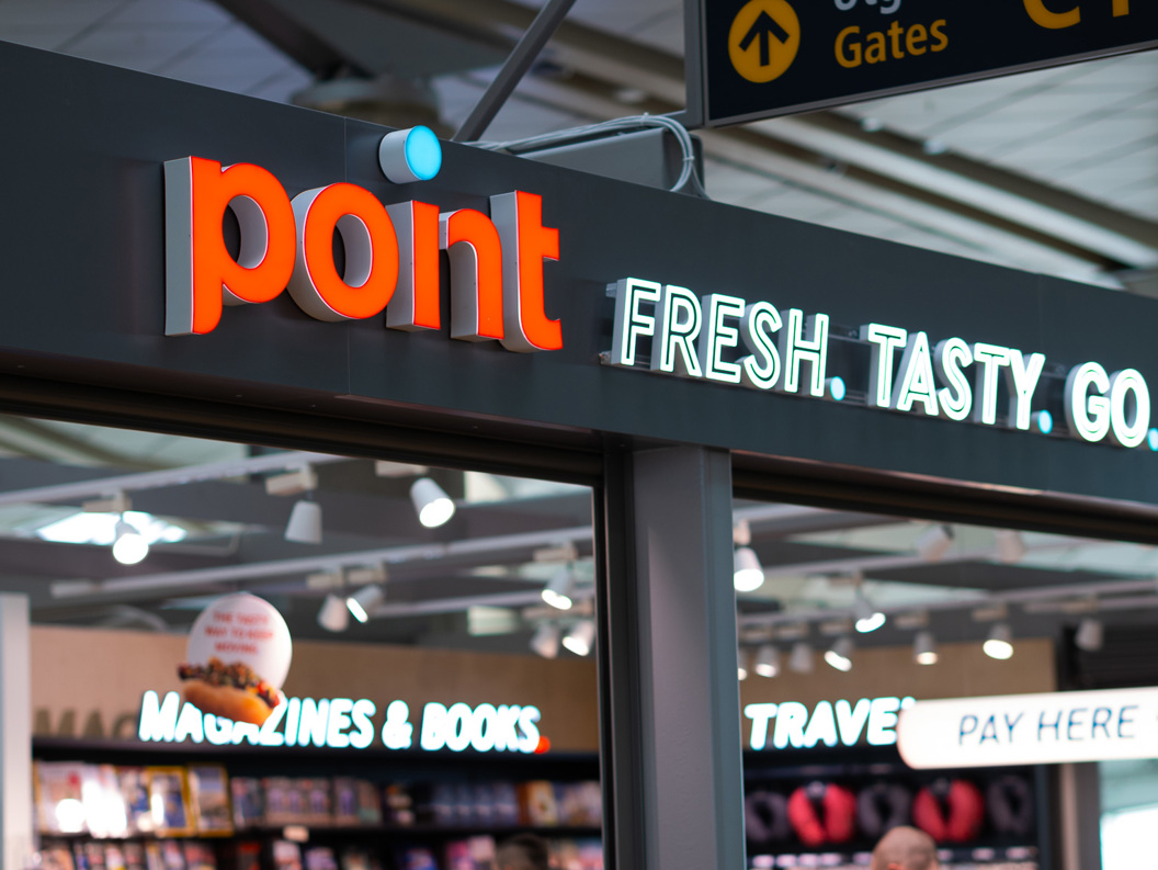 Butikkskilt på en flyplass med teksten "point" og "FRESH. TASTY. GO." vist over en butikk som tilbyr magasiner og bøker, med synlig takbelysning og skilt for reisemålporter