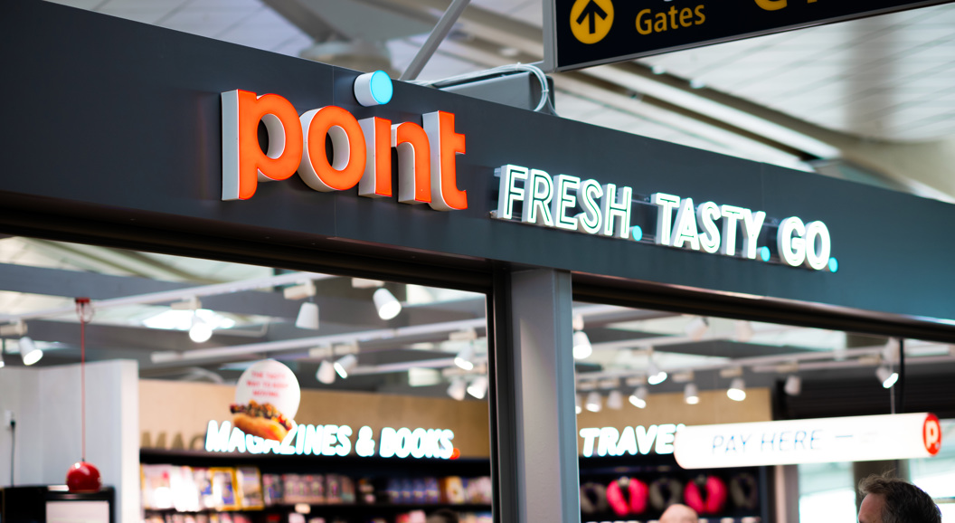 Skilt over butikk som viser teksten "point FRESH. TASTY. GO." i ein flyplassbutikk, med bokhandelseksjon i bakgrunnen