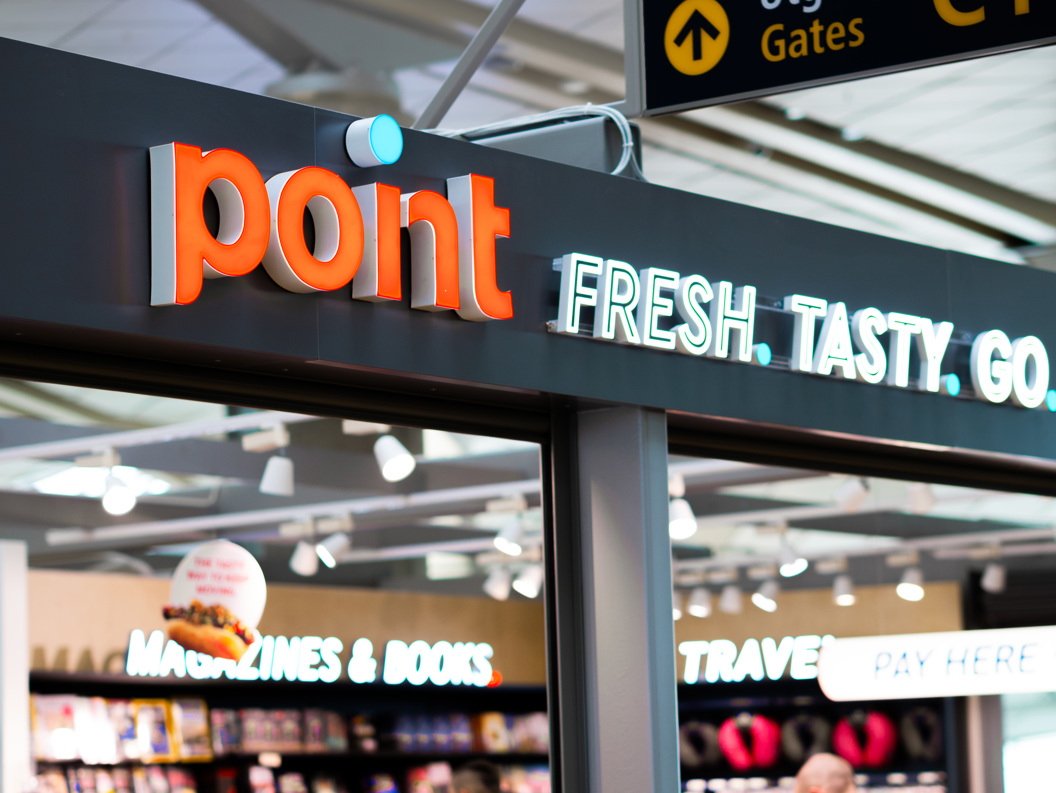 Skilt over butikk som viser teksten "point FRESH. TASTY. GO." i ein flyplassbutikk, med bokhandelseksjon i bakgrunnen
