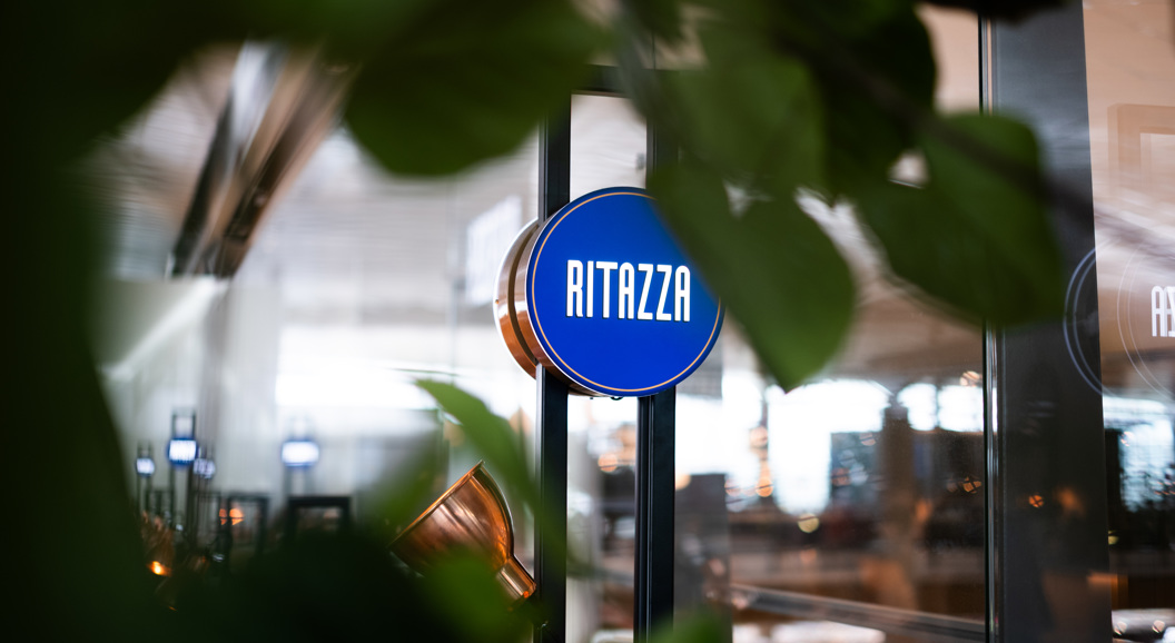 Skilt med teksten "Ritazza" på ein blå bakgrunn, omringa av grøne plantar i ein kafé