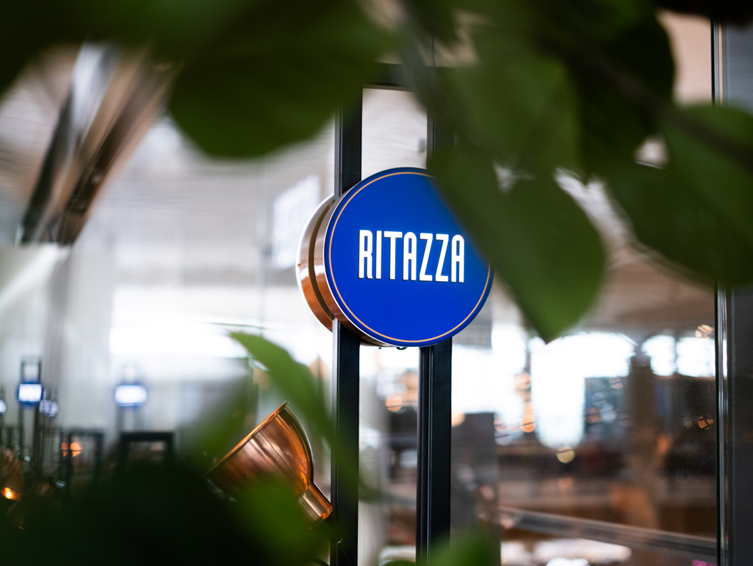Skilt med teksten "Ritazza" på ein blå bakgrunn, omringa av grøne plantar i ein kafé