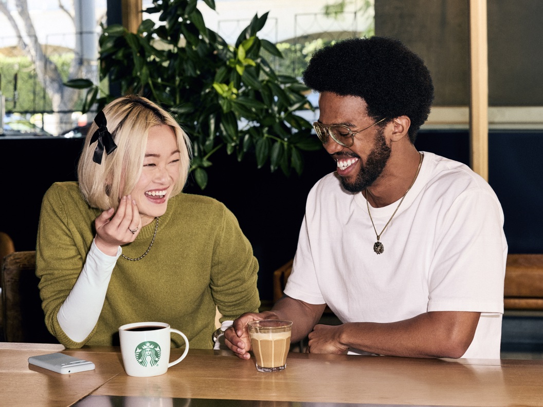 To personer ler og drikker kaffe ved et bord på en kafé. En kvinne har en grønn genser og en mann har en hvit t-skjorte. En Starbucks-logo vises på koppen
