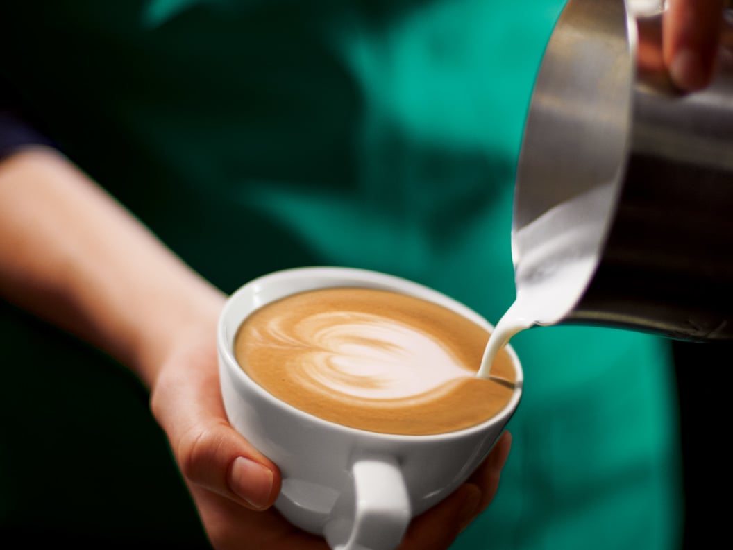 Barista pouring steamed milk into a cup of coffee creating a latte art pattern, held by a person in a green apron