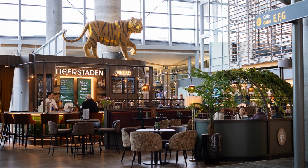 Tigerstadens vinbar på ein flyplass med ein stor tigerfigur på taket. Moderne interiør med sitteplassar og skilt for portar E, F, G i bakgrunnen