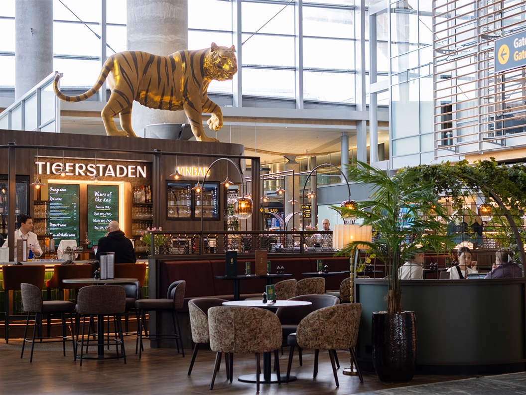 Moderne flyplassbar og loungeområde med sitteplasser, med en stor dekorativ tigerskulptur over "Tigerstaden Vinbar" skilt, med gjester og personale synlige, og et retningsskilt for porter E, F, G i bakgrunnen