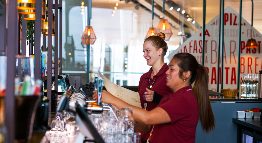To servitørar arbeider saman bak ein bar i ein restaurant, omgitt av glas og moderne belysning