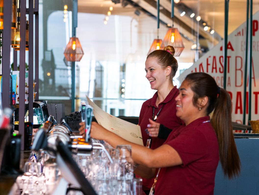 Bar staff working behind a modern bar counter, smiling and organizing drinks, with a stylish interior featuring hanging lamps and word art on the walls