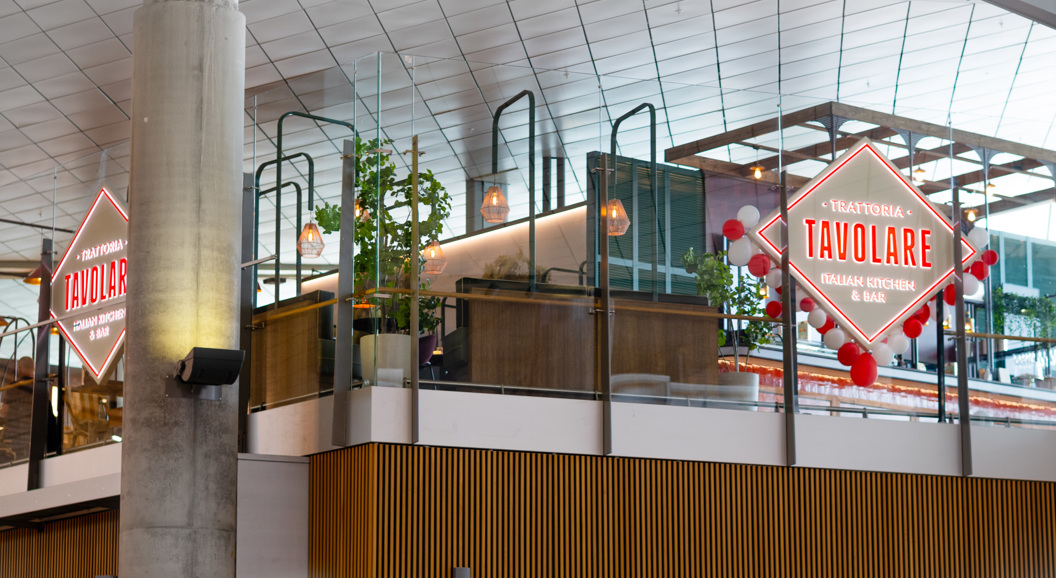 Restaurant Tavolare sin inngang med dekorativ lysreklame på ein flyplass, som viser moderne interiør og grønne planter