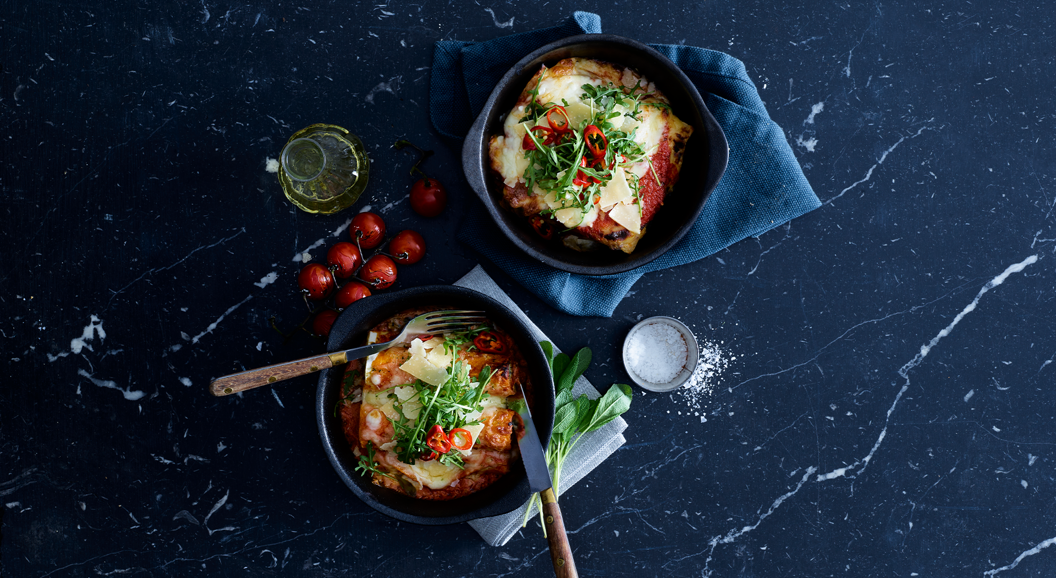 Two servings of lasagna in dark bowls on a marble surface, garnished with fresh greens and chili slices, accompanied by a small bottle of olive oil, cherry tomatoes, and a bowl of salt