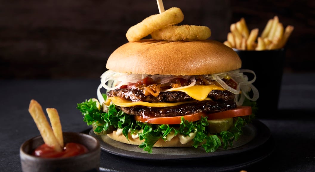 Delicious gourmet burger with lettuce, tomatoes, cheese, and onion rings on a bun, garnished with a skewer, accompanied by a side of fries and ketchup