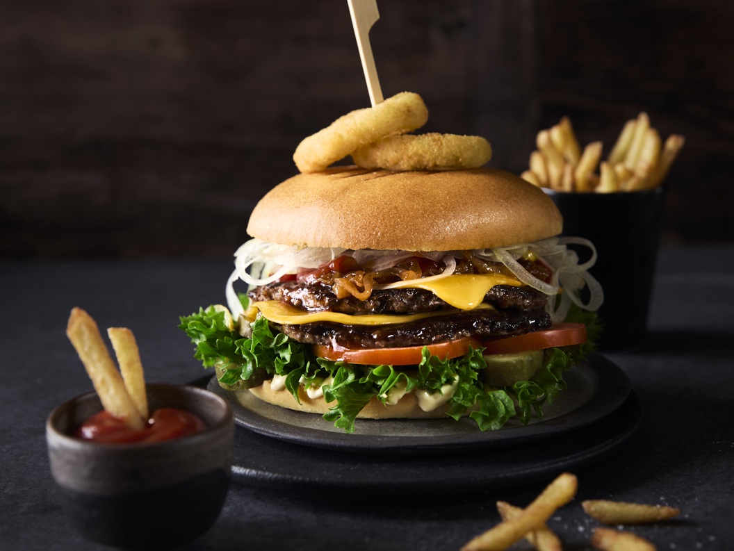 Saftig hamburger med tomat, salat, ost og løk, toppa med løkringar. Servert med pommes frites og ketchup i små skålar, på eit mørkt underlag