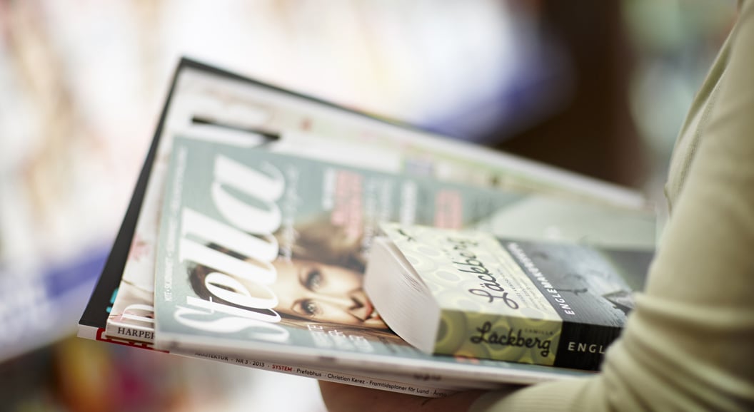 Person holding a stack of magazines and a book in a store, with the top magazine titled "Stella" and the book featuring an author's name