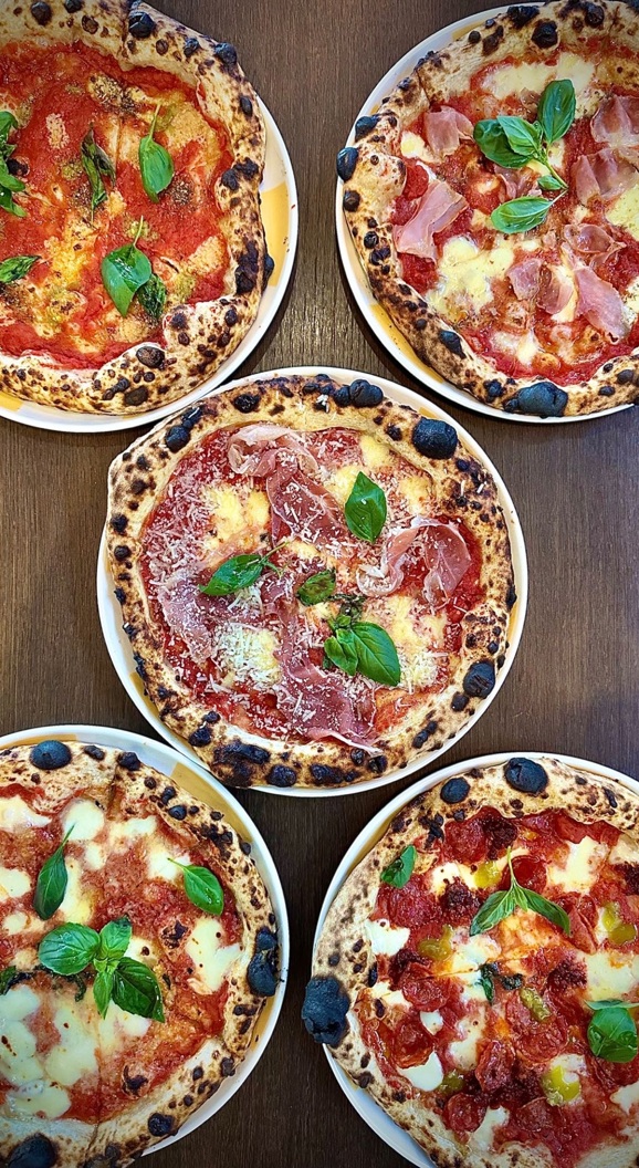 Five artisan pizzas with various toppings including basil, cheese, and cured meats on a wooden table