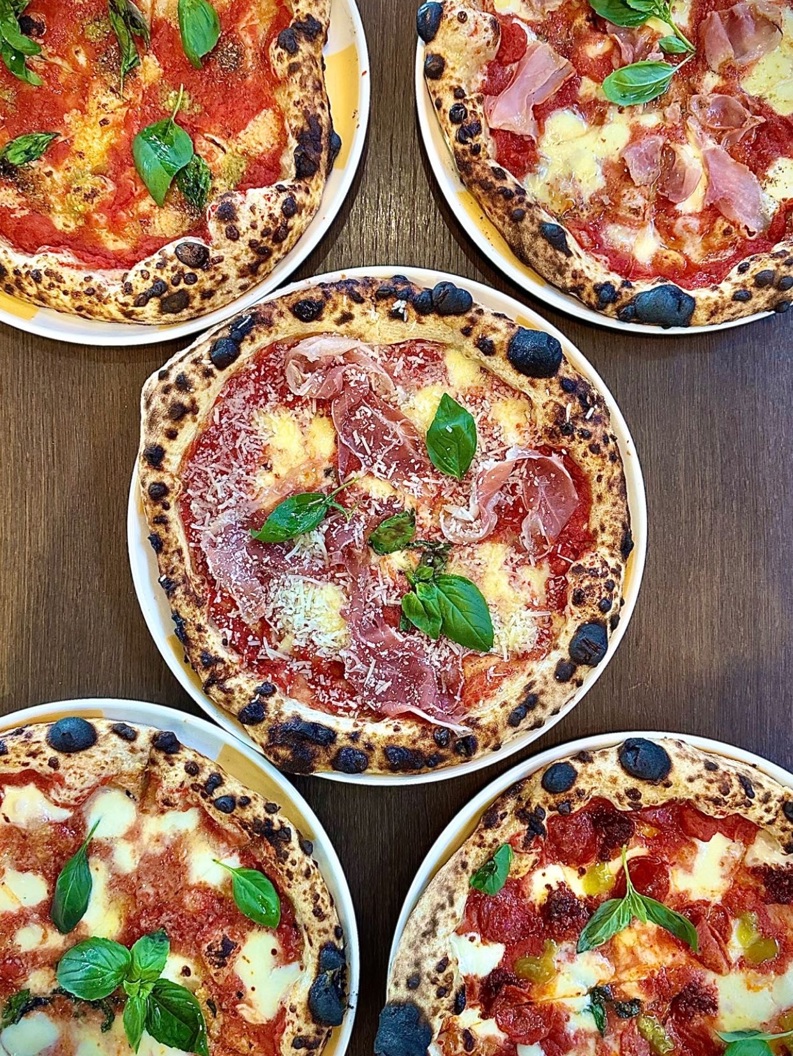 Five artisan pizzas with various toppings including basil, cheese, and cured meats on a wooden table