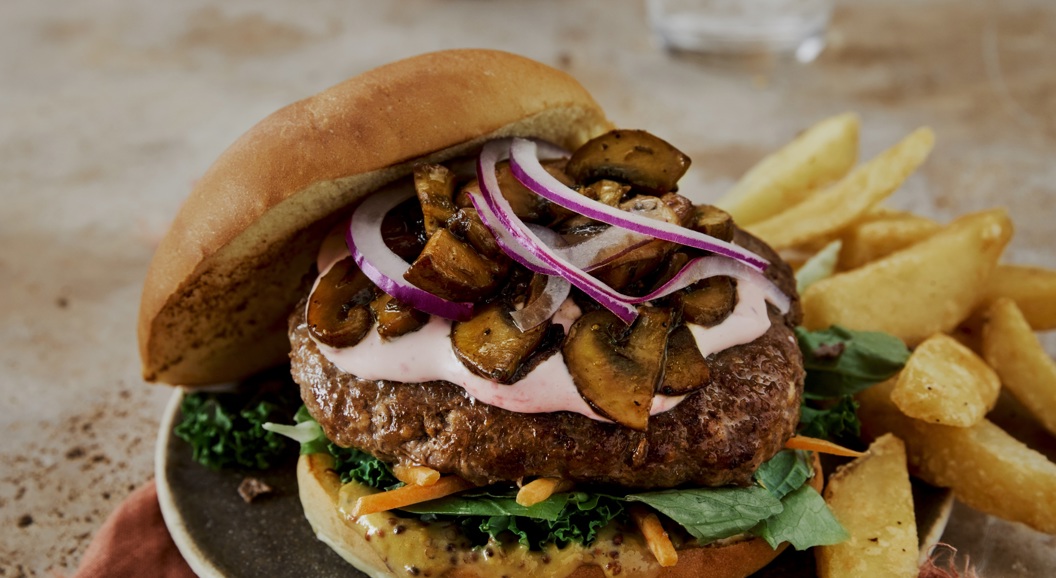 Saftig burger med rødløk, sopp, salat og dressing, servert med pommes frites på eit grått fat, ved sidan av glas med vatn