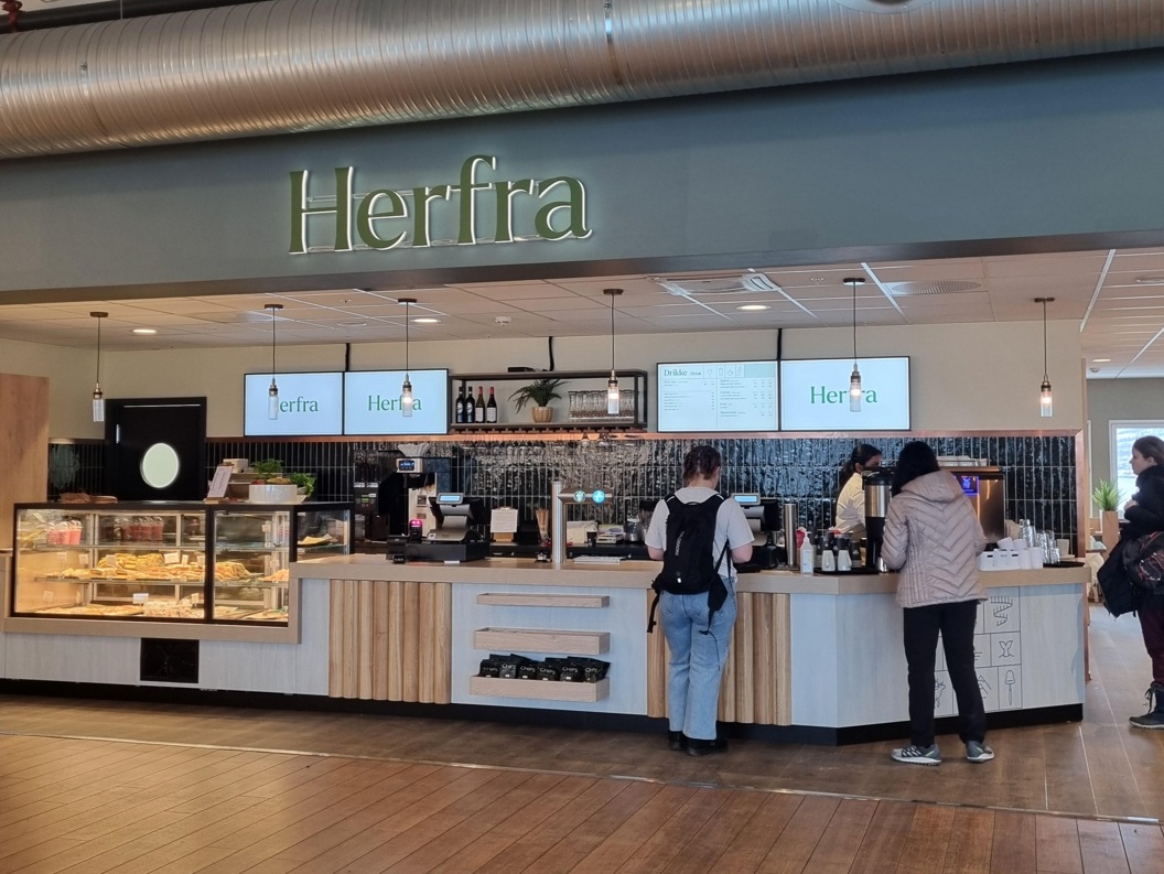 Interior view of a modern cafe named Herfra with customers ordering at the counter. The cafe features a display case with pastries, hanging lights, and digital menu screens above the counter