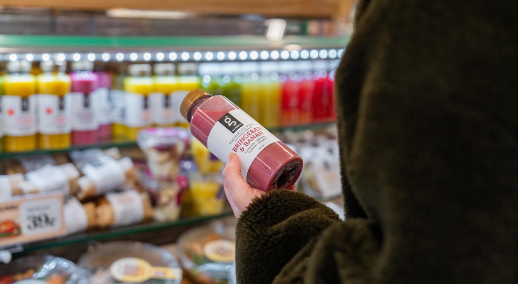 Person held raud smoothieflaske i butikk, med hyller full av juiceflasker i bakgrunnen