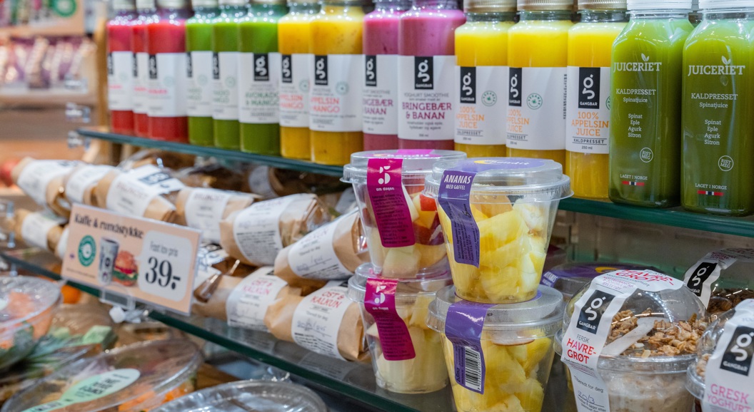 Drikkehyller med fargerike jusflasker og fersk mat, inkludert wraps, skåler med frukt og havregryn, i ein butikk