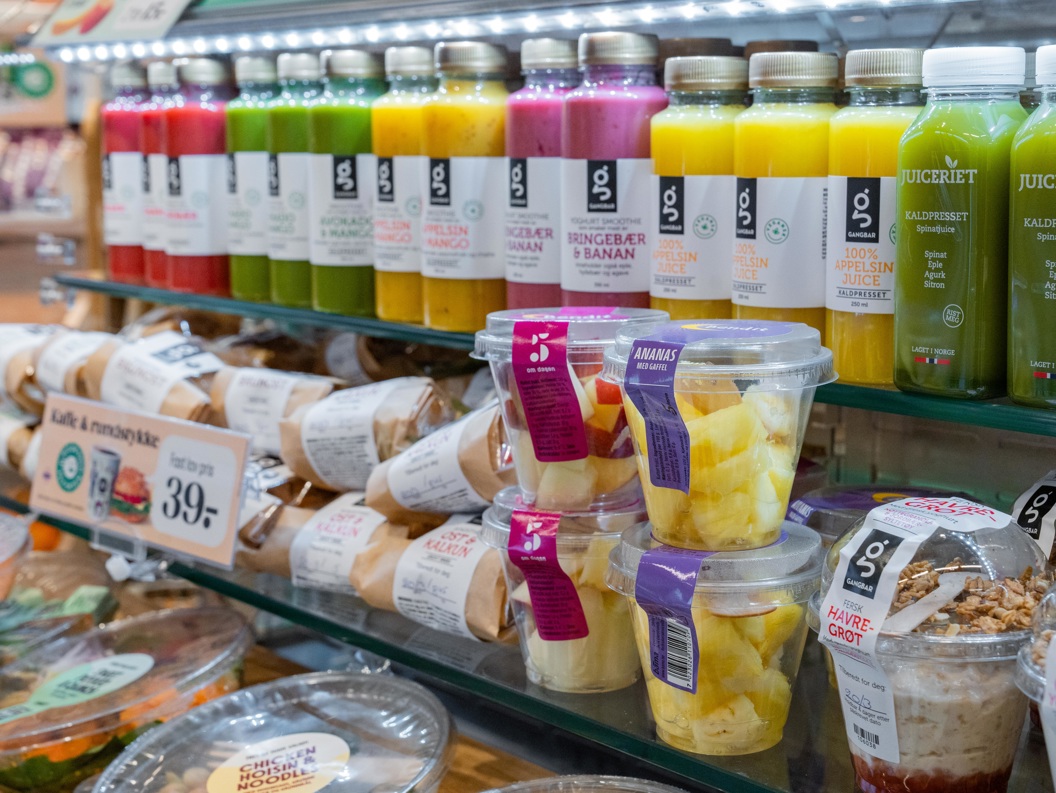 Supermarkedshylle som viser et utvalg av ferske juicer i fargerike flasker, delipakker og snackbeholdere med ananasbiter og granolayoghurt