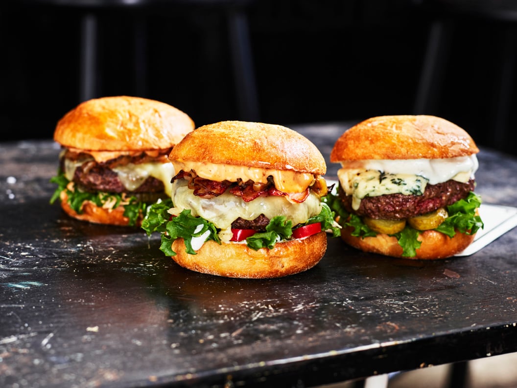 Three gourmet burgers with various toppings, including lettuce, cheese, pickles, and bacon, on a rustic black table