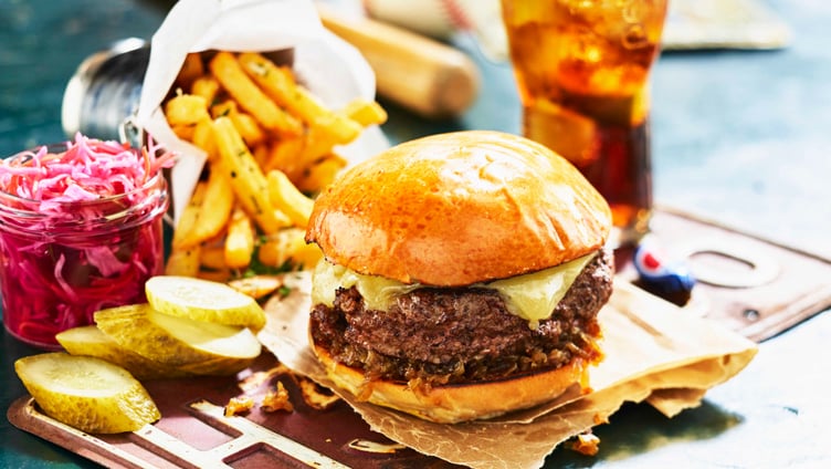 Juicy cheeseburger with melted cheese, accompanied by crispy French fries, sliced pickles, and coleslaw, with a glass of iced cola on a rustic table setting