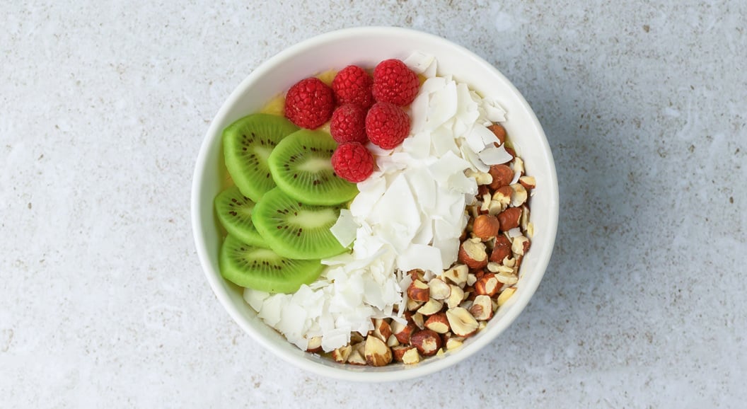 Healthy breakfast bowl with sliced kiwi, raspberries, coconut flakes, and chopped nuts on a light granite background